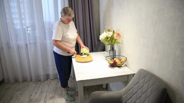 Older woman chopping fresh green cabbage on wooden cutting board with knife in kitchen for healthy meal preparation, showing everyday domestic home cooking activity.