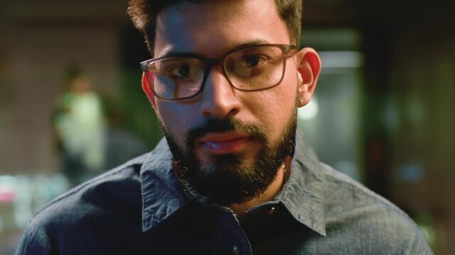 Indian business man working at night in office computer lights reflection in glasses. Arabian ethnic businessman male guy programmer trader wear eyeglasses trading coding looking at camera at evening