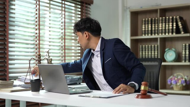 Asian lawyer working in a law firm, multitasking with a laptop, signing documents and talking on a smartphone while sitting at a desk with a gavel and scales of justice