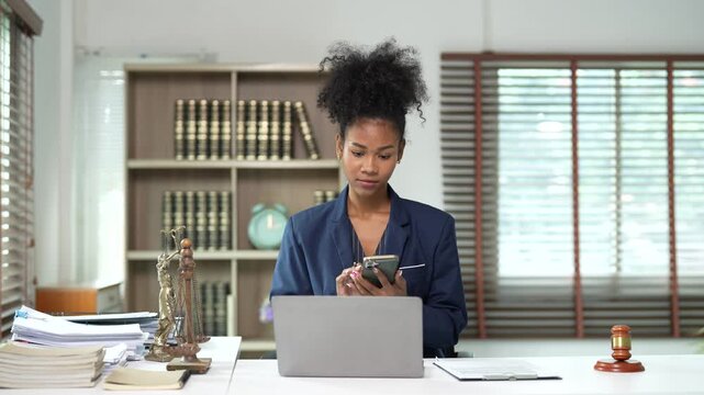 African american female lawyer working on laptop and legal documents at desk in office, representing justice with the scales of justice and a judge's gavel nearby