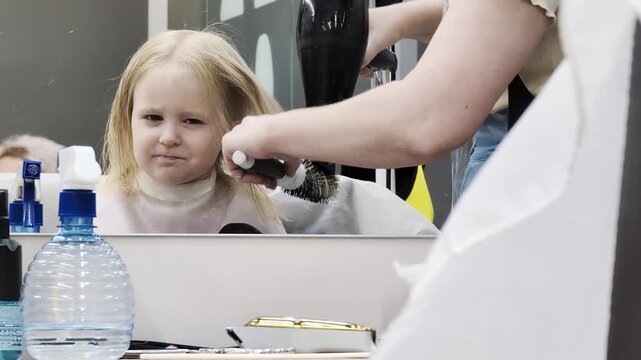 Little girl receives professional hair styling service at hair salon, hairdresser drying blonde hair of child with brush and hairdryer, reflection of child face