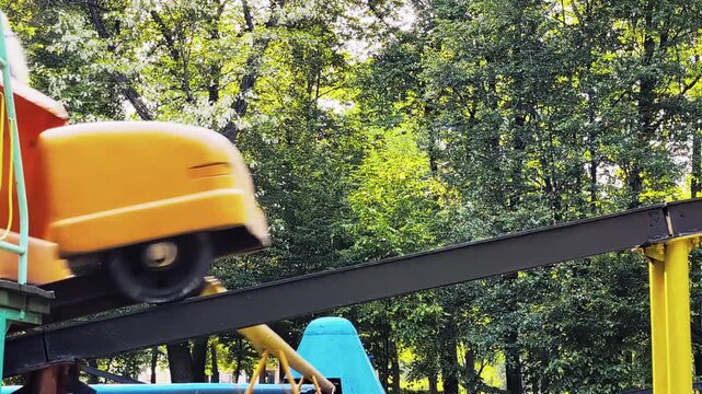 Colorful roller coaster cars moving fast on elevated tracks against lush green trees in amusement park, capturing excitement of public park attraction.