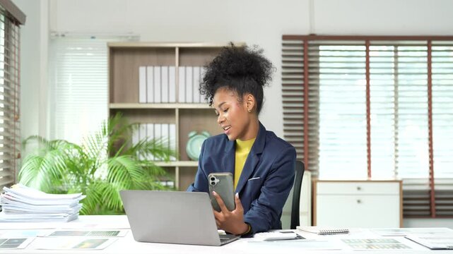 Young african american businesswoman talking on a video call using her smartphone while holding a coffee cup, showing a wide range of emotions in her bright modern office