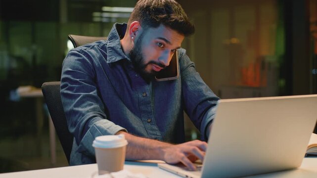 Busy multitasking Arabian business man emotional talking phone working with laptop at evening office Indian guy stressed overworked Hindu businessman male talk call looking in computer and textbook