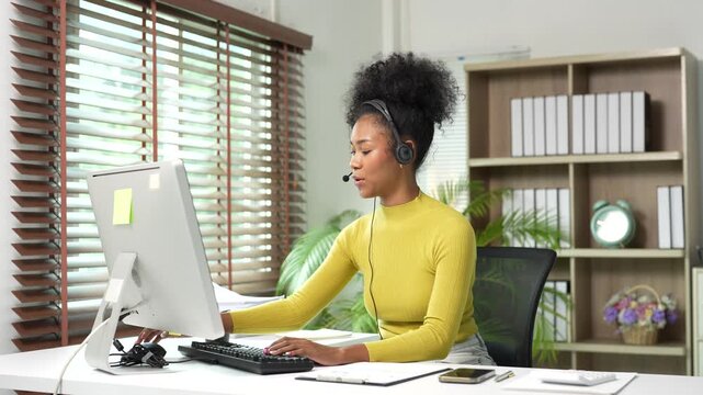 African American call center agent with a headset providing customer service and consulting online while working at her desk in a modern office with natural light