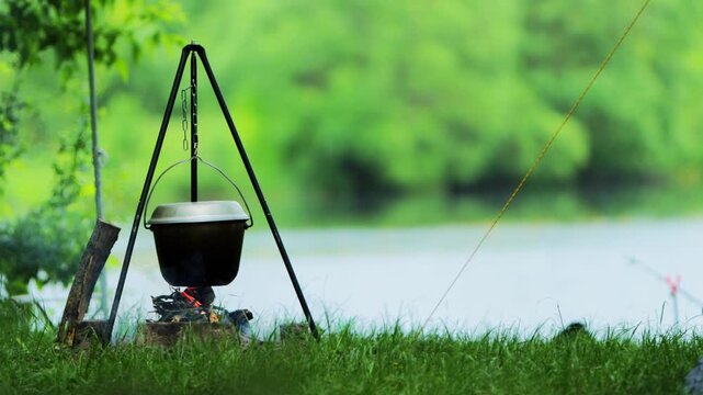 Cooking pot suspended over campfire by tripod on grassy bank near serene lake with lush green foliage background, illustrating outdoor recreation and peaceful nature, copy space