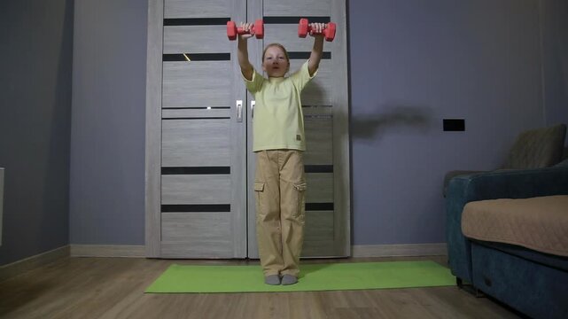Young girl performing shoulder press exercise with dumbbells in room. Child fitness workout indoors, strength training activity at home, health and wellness.