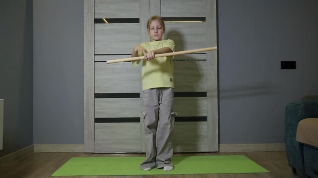 Little girl performs exercises with wooden stick indoors on yoga mat, engaging in physical activity for development of body and health, demonstrating focus and discipline.