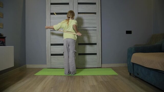 Young girl performing back stretches with wooden stick at home, engaging in fitness activity on yoga mat indoors, focused on health and physical well-being.