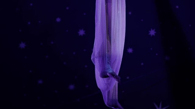 Female performer aerial acrobatics on purple silk fabric against dark starry background circus stage performance graceful movement of body and fabric slow mo