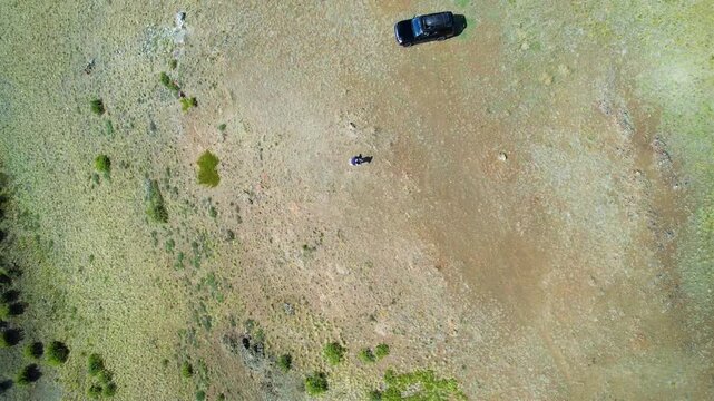 High overhead drone view of two young people embracing on wide grassy plateau beside parked SUV, with strong midday shadows, open wilderness texture, and remote mountain setting