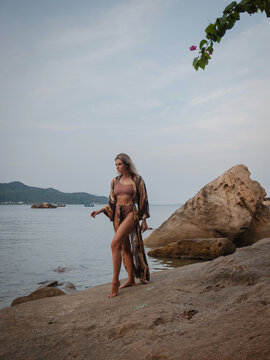 Fashion model in patterned beach kimono and bikini standing on tropical rocky shoreline by calm ocean