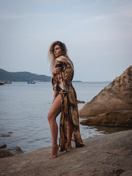 Fashion model wearing abstract print beach kimono posing on rocky tropical shoreline by calm ocean