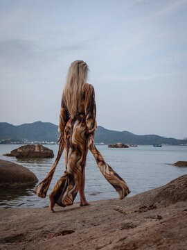 Woman in flowing abstract print beach kimono enjoying peaceful moment on rocky tropical shoreline