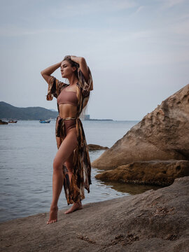 Fashion model in patterned beach kimono and bikini standing on tropical rocky shoreline by calm ocean