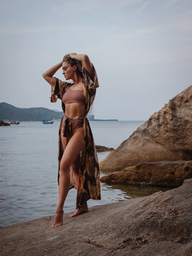 Fashion model in patterned beach kimono and bikini standing on tropical rocky shoreline by calm ocean