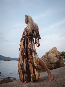 Fashion model wearing abstract print beach kimono posing on rocky tropical shoreline by calm ocean