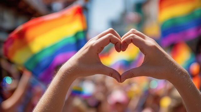 A person forms a heart gesture with hands against a background of waving rainbow flags. Concept of universal love, support for LGBT rights, and diversity.