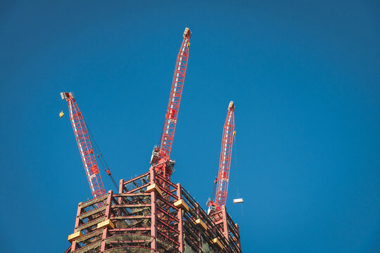 Red luffing jib cranes lift heavy materials on rooftop of skyscraper structure in Taipei, Taiwan, showing under construction mechanisim in modern high-rise building featuring steel braced skeleton
