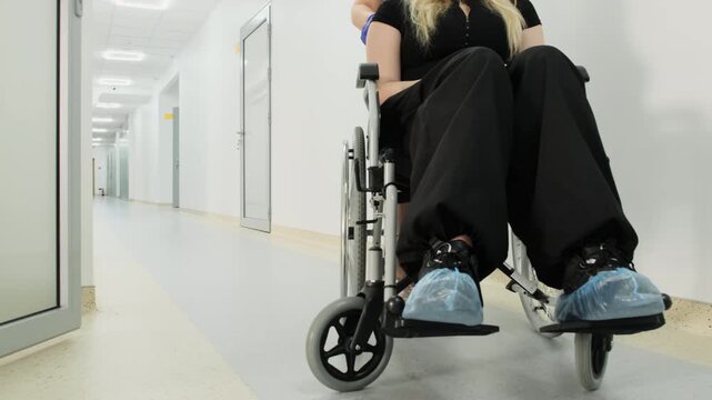 Attentive nurse carefully wheels woman out of procedure room. Doctor guides patient through hospital corridor toward hospital ward. Slow motion