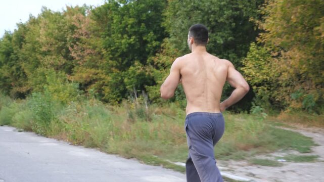 Rear view of muscular man with wireless headphones jogging along trail near forest at early autumn. Athletic guy running along road at nature. Young sportsman training outdoor. Healthy lifestyle