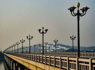 Obraz premium Long concrete bridge with repeating ornate street lamps crosses water. Buildings visible on distant tree covered hill. Daytime scene with clear sky.