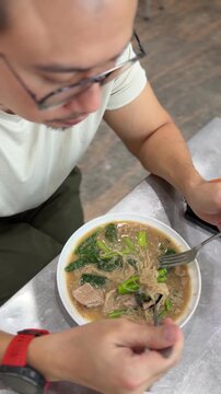 Asian Man Enjoying Authentic Thai Noodles with Pork and Green Vegetables at a Local Eatery