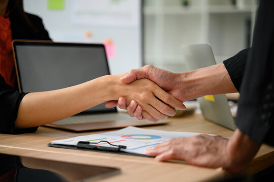 Close Up Business Handshake Between Colleagues Across Office Desk with Laptop and Financial Charts