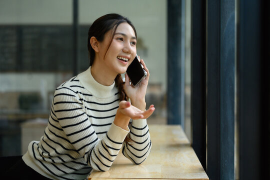 Happy Young Asian Woman Talking on Smartphone by Window in Cafe, Smiling and Gesturing