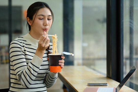 Young Asian Woman Eating Instant Noodles from Cup at Office Desk with Laptop, Quick Convenient Meal Break
