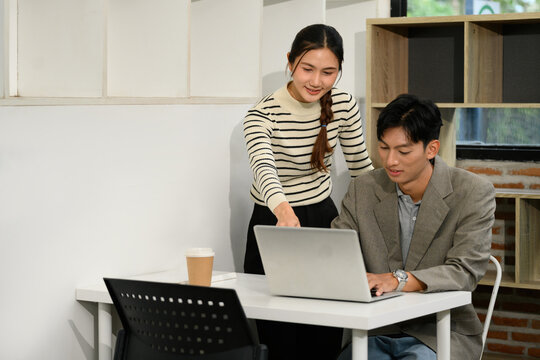 Two Asian Colleagues Working Together on Laptop at Modern Office Desk with Coffee Cup