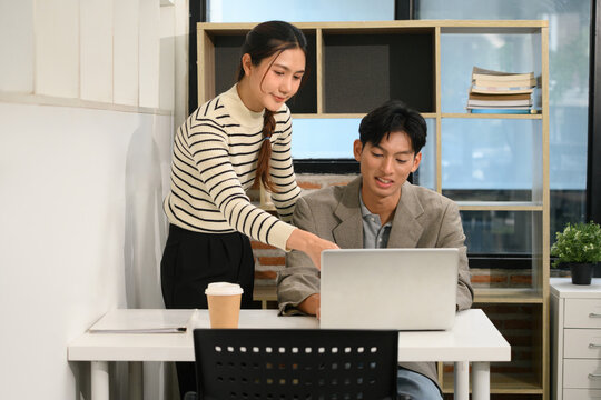Two Asian Colleagues Working Together on Laptop in Modern Office, Woman Pointing at Screen While Man Sits at Desk