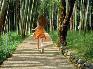 Naklejka premium Woman running on a gravel path through a bamboo forest, back view in an orange skirt, summer sunlight filtering between trunks, motion and freedom on a peaceful park walkway.
