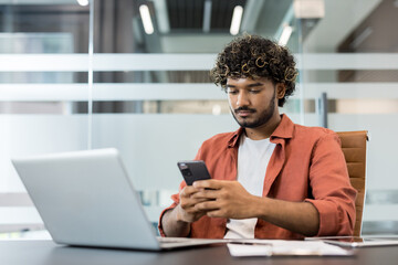 Obraz premium Indian office worker looking at his smartphone while sitting at a desk with a laptop, reflecting modern workplace distractions and the challenge of multitasking between devices