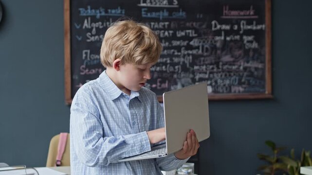 Portrait of Caucasian boy typing on laptop in classroom and looking at camera. Video is suitable for promoting STEM education, coding bootcamps, or modern school programs
