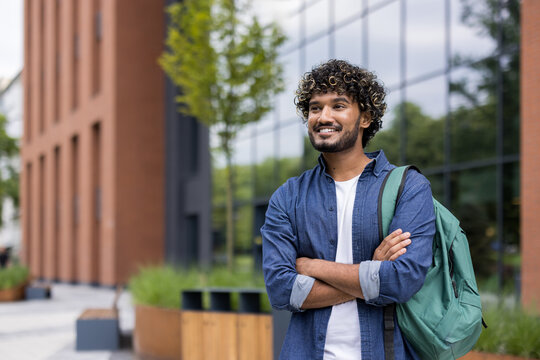 Young man student with a backpack smiling and looking away, arms crossed, standing on a modern university campus building background