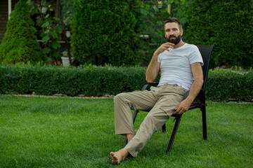 Fototapeta premium Confident bearded man sitting on outdoor chair in a green backyard garden, looking at camera with a thoughtful expression. Relaxed lifestyle, leisure time, personal freedom, mindfulness