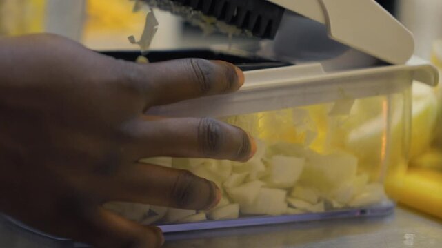 A close-up shot of a chef's hands in a professional kitchen using a manual vegetable chopper to efficiently dice fresh onions into a clear container