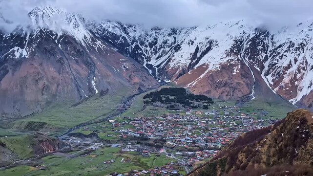Snow-covered peaks of the Caucasus Mountains rise above a sprawling village in Stepantsminda, Georgia under an overcast sky.