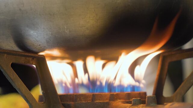 A detailed close-up shot of an industrial gas stove burner with bright blue and orange flames heating the bottom of a large metal pot in a professional kitchen setting.