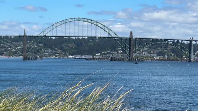 The historic steel arch Yaquina Bay Bridge stretches across calm harbor waters, viewed from a rocky shoreline in Newport, Oregon.