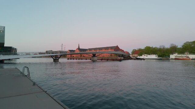 City river sunset bridge buildings, gimbal stabilizer shot. 60fps cinematic push in walking on Copenhagen bank looking at bikes on overpass and moored boats.
