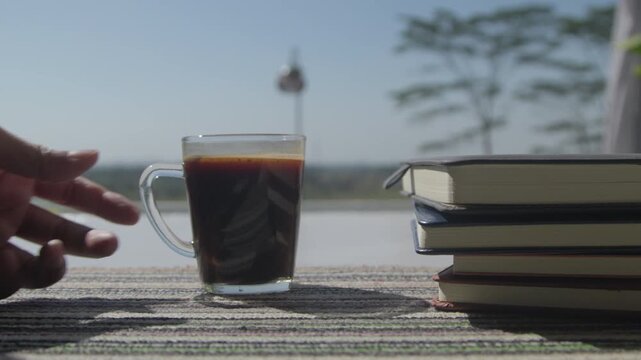 A hand place a cup of black coffee beside a neat stack of books on a carpet bathed in morning sunlight, creating a calm reading and relaxation scene at home