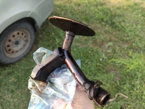 Hand holding oil pump pick up tube with strainer, bottom view, car in background