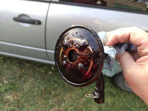 Hand holding dirty oil pump strainer with sludge and bubbles, car in background