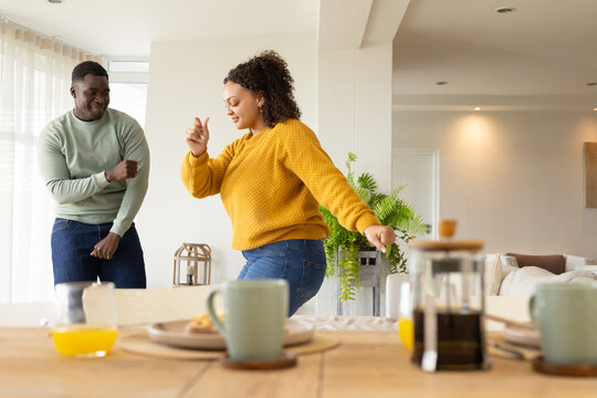 African American couple dancing near dining table in home with French press, mustard sweater