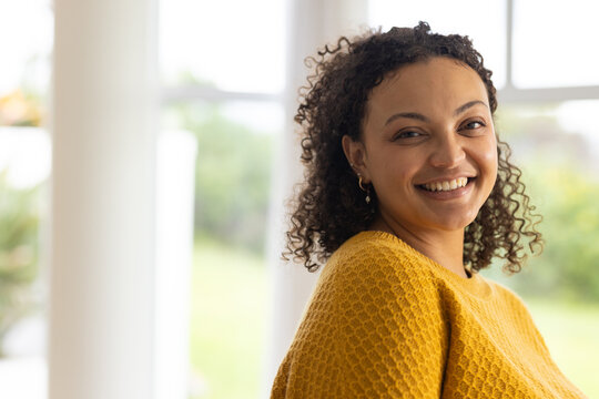 African American woman smiling, looking over left shoulder by window frames wearing mustard sweater
