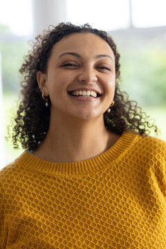 Woman smiling tilting head wearing mustard sweater gold hoop earrings with pearls near large window