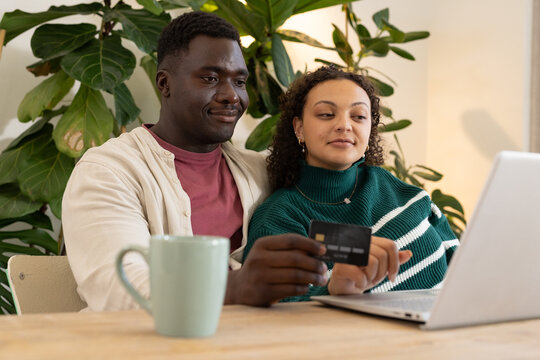 Diverse couple sitting at wooden dining table using laptop, holding credit card and mug