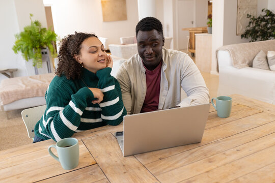 African American couple sitting at dining table, woman in green sweater, viewing laptop with mugs